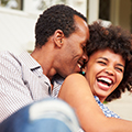 Couple métisse avec un homme chuchottant à l'oreille d'une femme souriante pour symboliser le jeu érotique de la langue fourchée - Image de l'article Conseils pour s'éclater au lit et s'amuser lors du rapport sexuel - Rubrique Jouer à des jeux sexuels pour booster le désir de l'autre et pimenter ses ébats