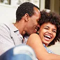 Couple métisse avec un homme chuchottant à l'oreille d'une femme souriante pour symboliser le jeu érotique de la langue fourchée - Image de l'article Conseils pour s'éclater au lit et s'amuser lors du rapport sexuel - Rubrique Jouer à des jeux sexuels pour booster le désir de l'autre et pimenter ses ébats