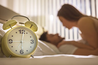 Réveil au premier plan et couple au second, pour symboliser le besoin de prendre son temps pendant les moments intimes - Comment satisfaire les femmes au lit