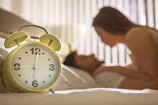Réveil au premier plan et couple au second, pour symboliser le besoin de prendre son temps pendant les moments intimes - Comment satisfaire les femmes au lit